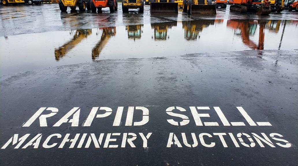 Rapid Sell machinery auctions sign painted on wet pavement at equipment yard